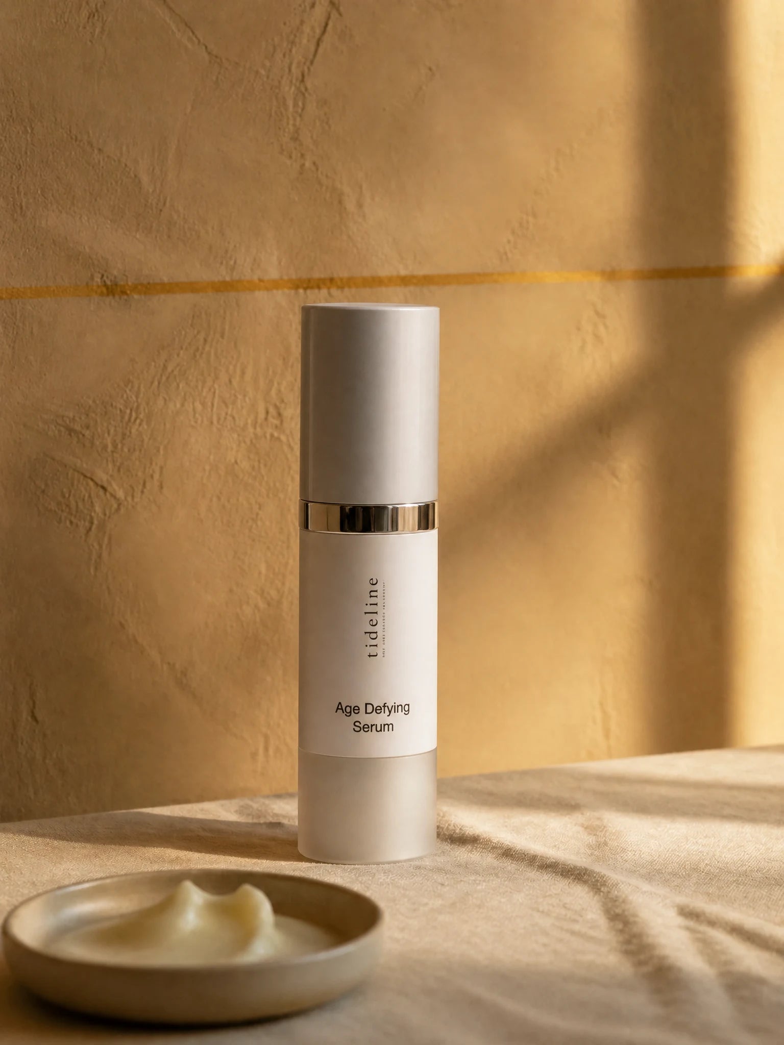 Tideline Age-Defying Serum, a white airless pump bottle on warm linen in soft window light