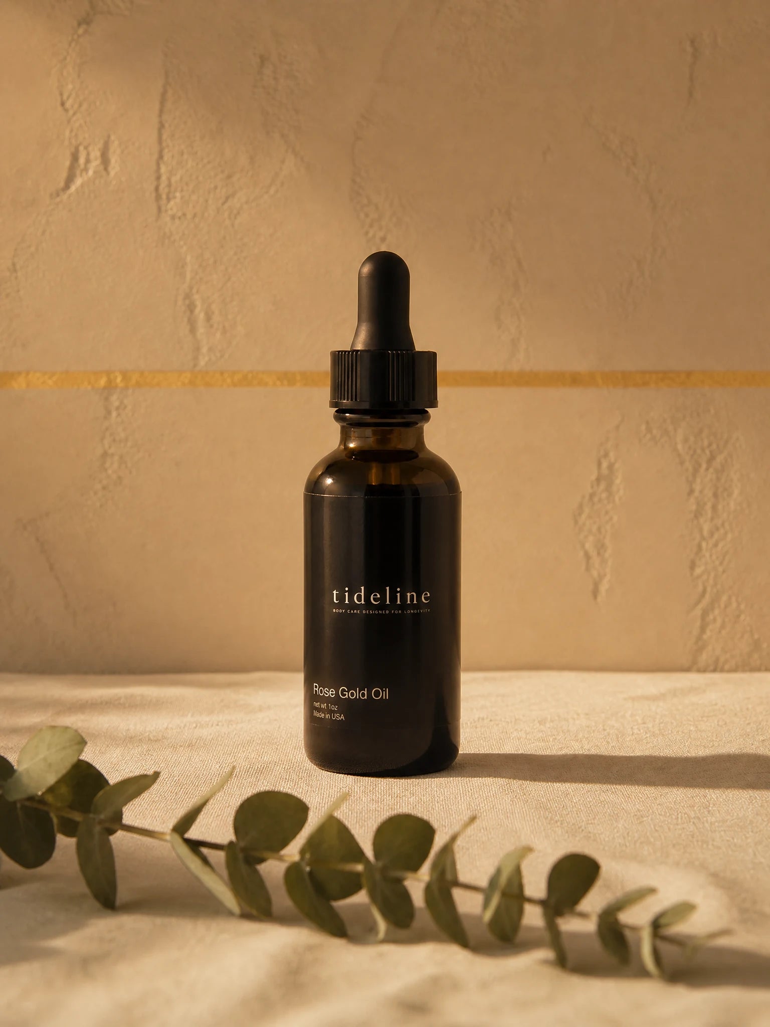 Tideline Rose Gold Oil, a black glass dropper bottle on warm linen with eucalyptus